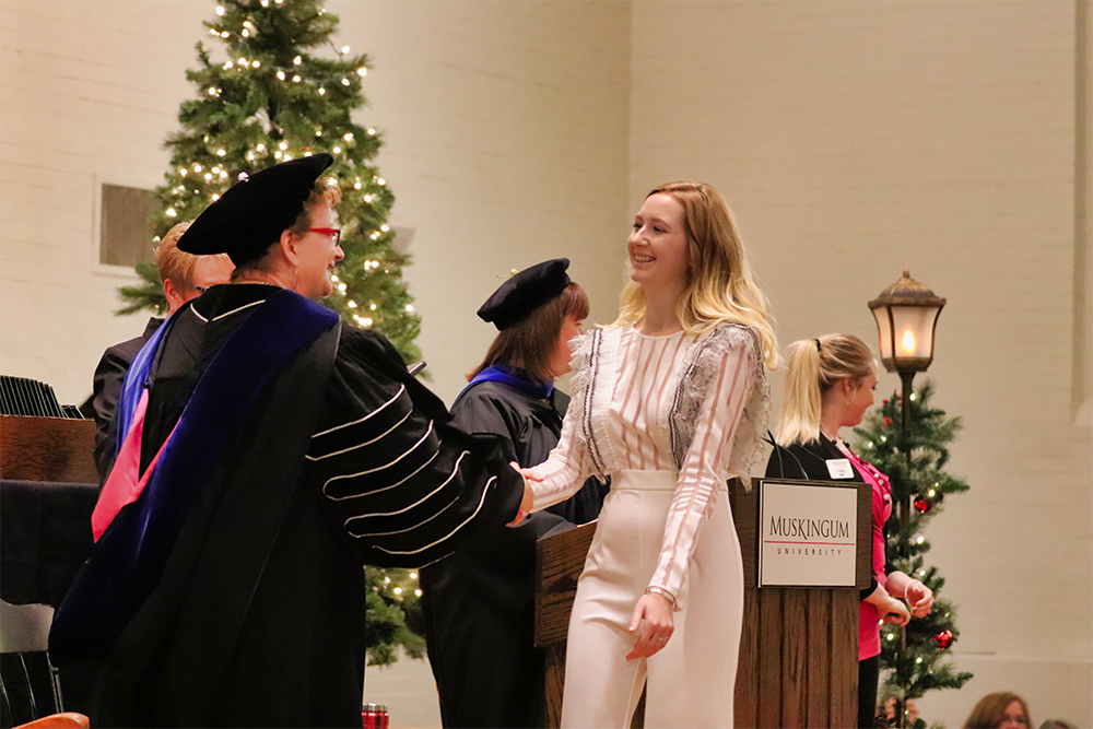 a blonde female student dressed in white crossing the stage and shaking hands with President Hasseler