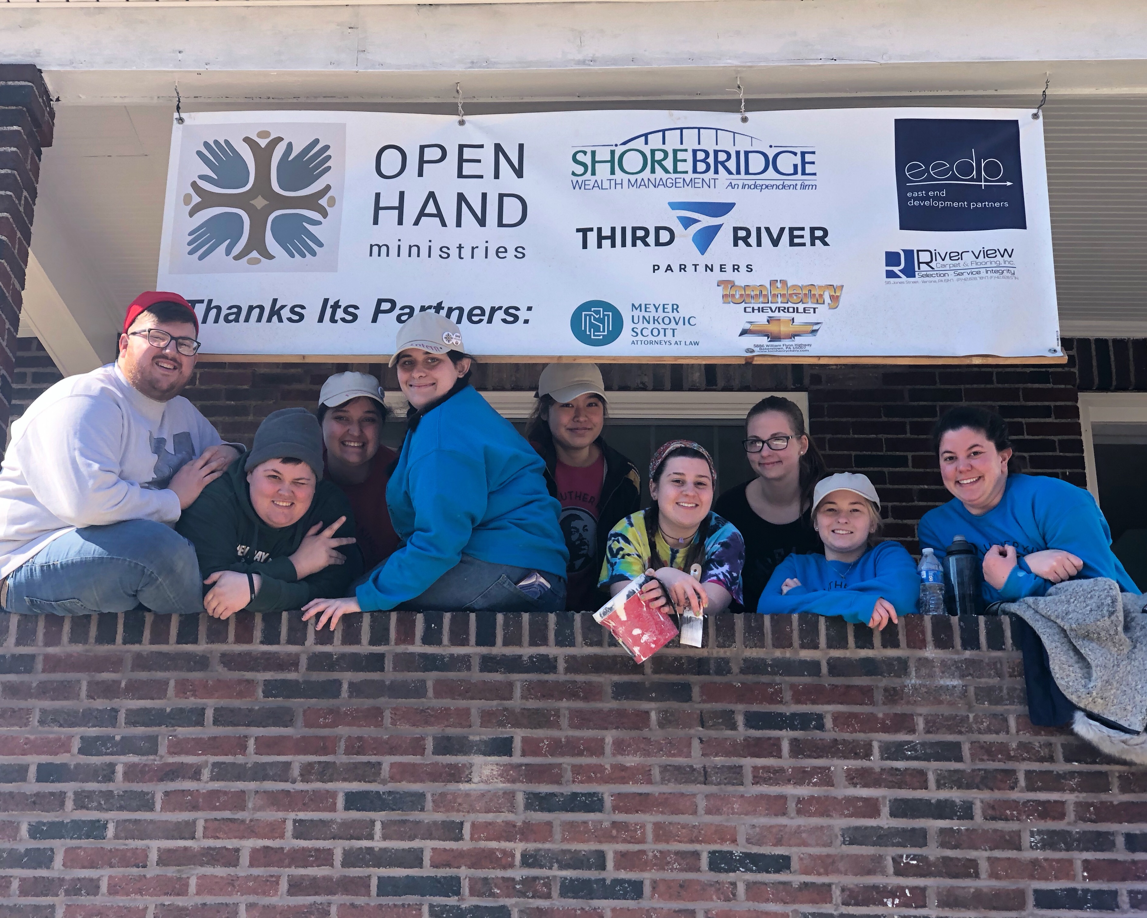 Students in bright blue sweatshirts smiling on a brick porch under a banner listing Open Hands Ministries and other sponsoring organizations in Pittsburgh PA.