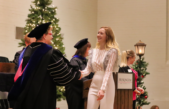 a blonde female student dressed in white crossing the stage and shaking hands with President Hasseler