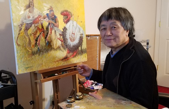 Professor Yan Sun sitting in front of one of his paitings of Native Americans holding his brush and pallette