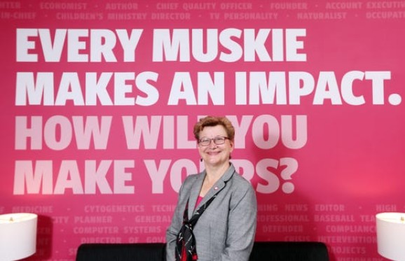 susan hasseler posing in front of a wall with the text "every muskie makes and impact.  How will you make yours?"