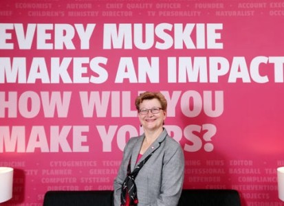 susan hasseler posing in front of a wall with the text "every muskie makes and impact.  How will you make yours?"